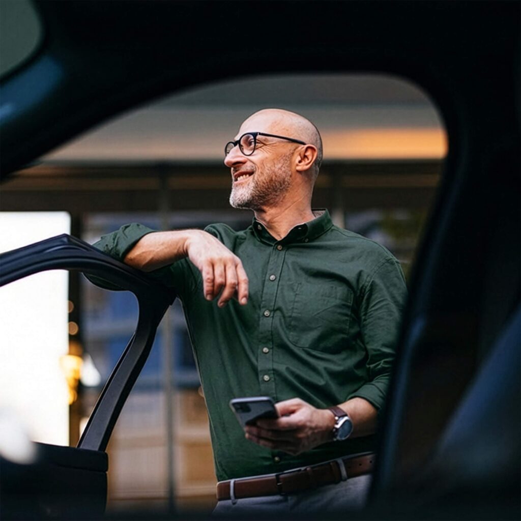 Henkilö seisoo autoa vasten puhelin kädessä ja katsoo kauas.